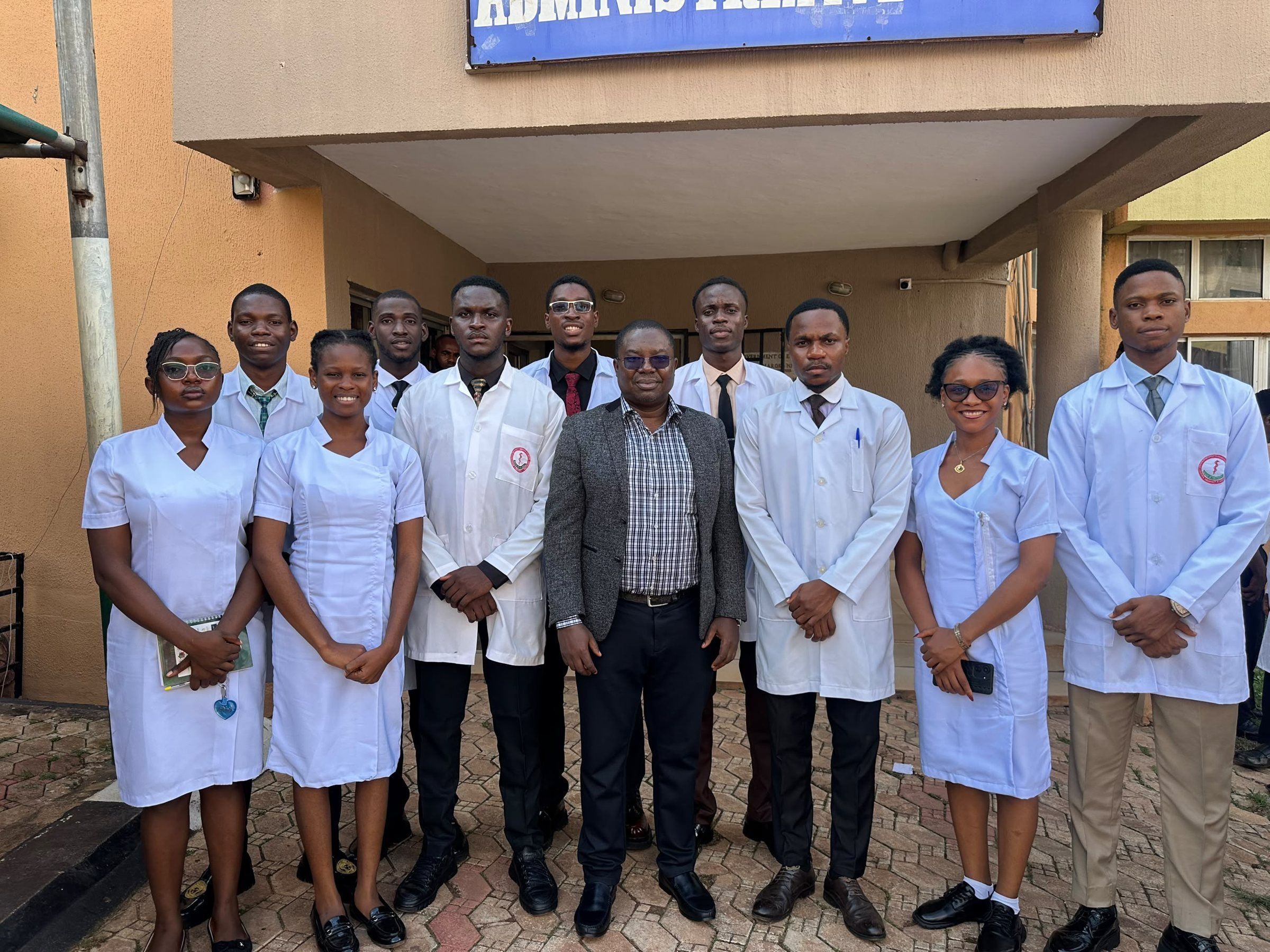Ten ESUMSA executives in white coats and uniforms standing with a staff member outside the Administrative Block.
