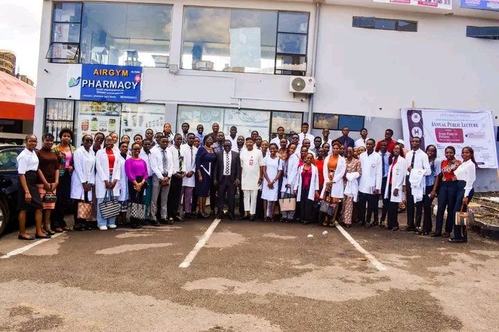 A large group of students, many in white coats, standing outside beside an annual public lecture banner.