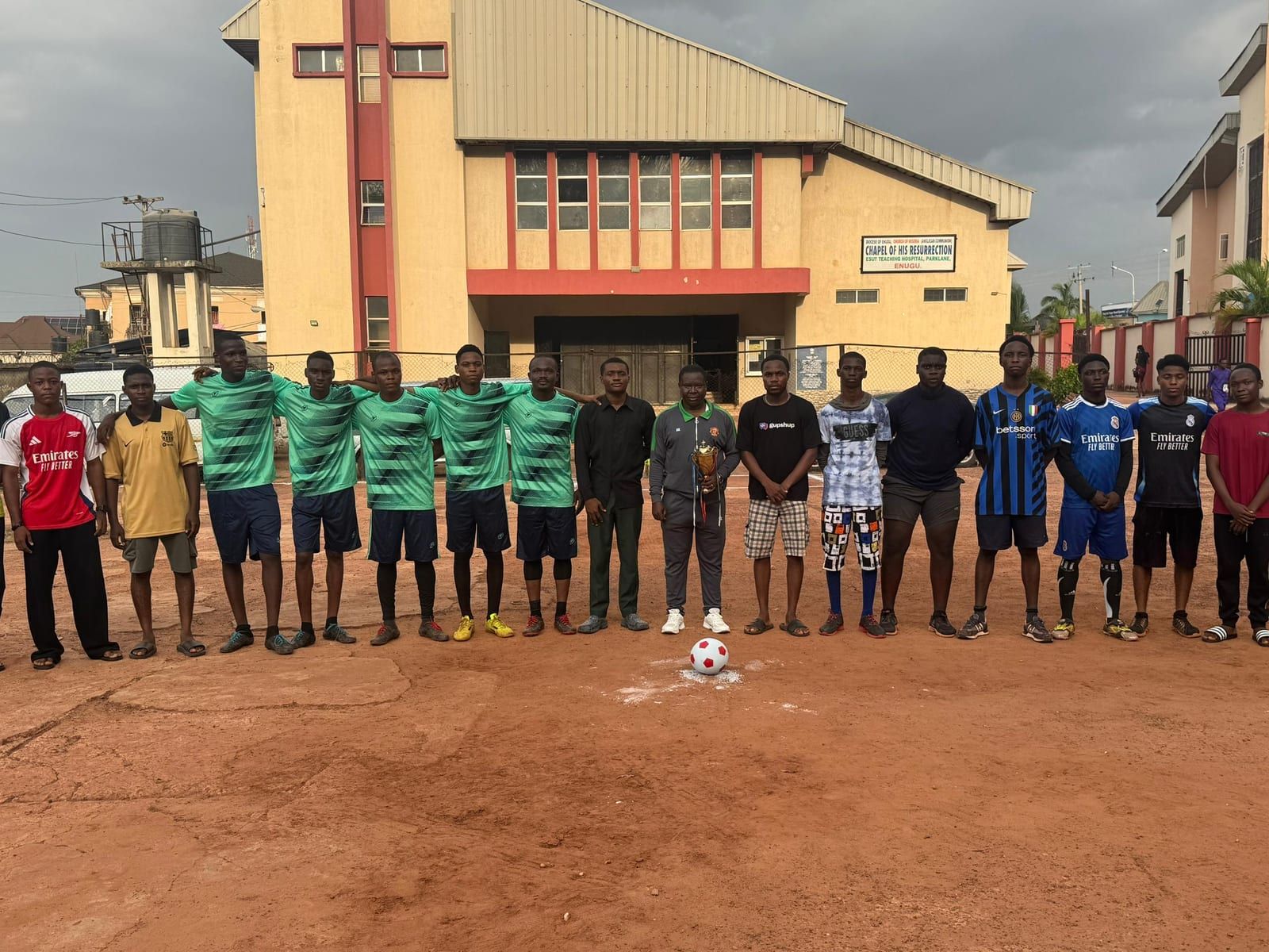 Two football teams, one in green and one in assorted kits, lined up in front of a college building.